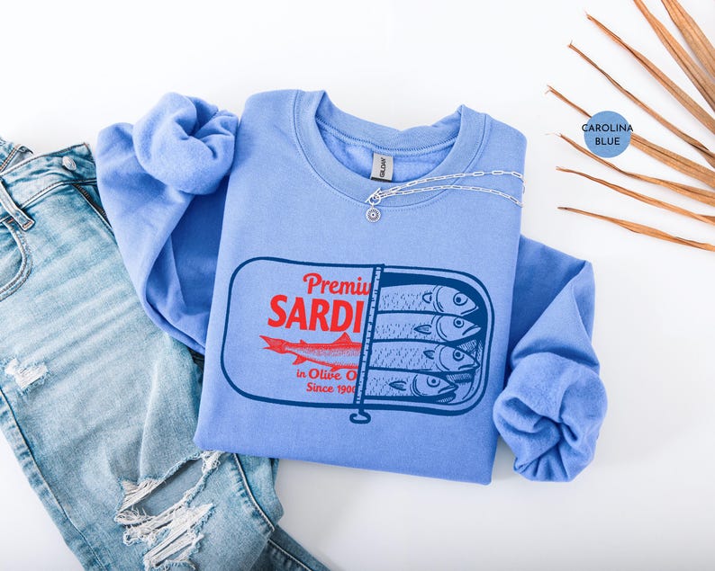 May include: Carolina blue sweatshirt with a sardine can graphic. The graphic features the text "Premium SARDI" in red, a red fish, and the text "in Olive Oil Since 1900" in black. A silver necklace is draped across the sweatshirt.