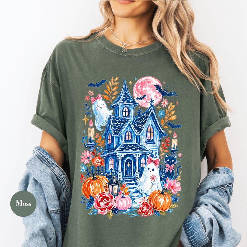 K&ouml;nnte beinhalten: Ein moosgr&uuml;nes T-Shirt mit einem farbenfrohen Grafikdruck eines blauen Spukhauses, zwei Geistern mit rosa Schleifen, orangefarbenen K&uuml;rbissen, roten und rosa Blumen, einer schwarzen Katze, Flederm&auml;usen und einem gro&szlig;en rosa Mond.