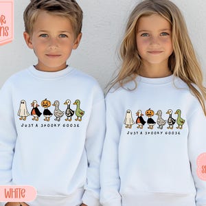 May include: Two children wearing white sweatshirts with a Halloween design. The design features cartoon geese dressed as a ghost, vampire, pumpkin, and mummy. The text "JUST A SPOOKY GOOSE" is printed below the geese.