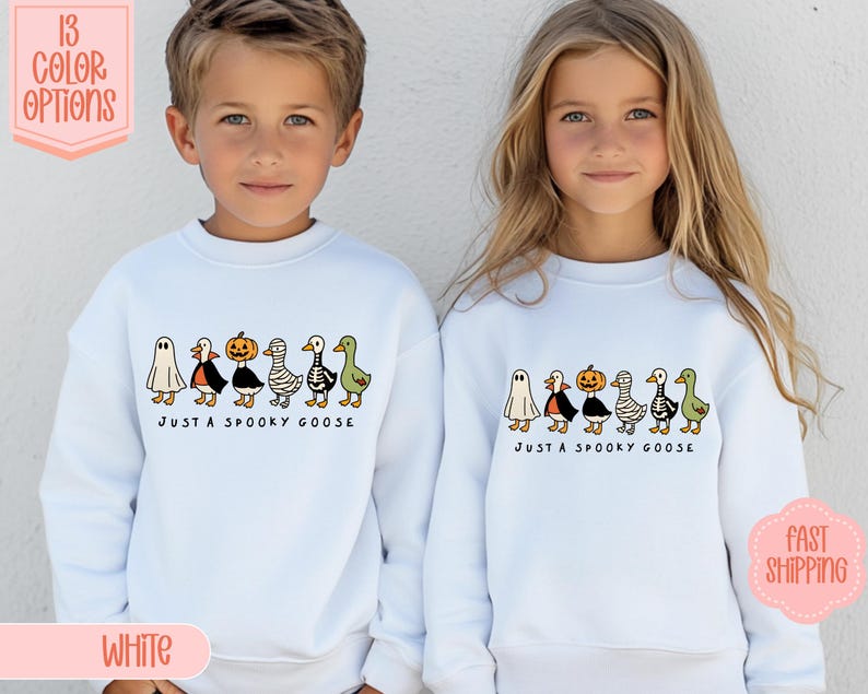 May include: Two children wearing white sweatshirts with a Halloween design. The design features cartoon geese dressed as a ghost, vampire, pumpkin, and mummy. The text "JUST A SPOOKY GOOSE" is printed below the geese.