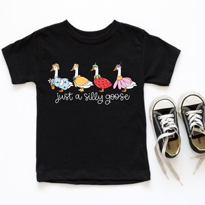 以下が含まれることがあります： 黒い半袖Tシャツに、異なる衣装を着た5羽のガチョウと「Just a silly goose」というテキストのグラフィックがプリントされています。