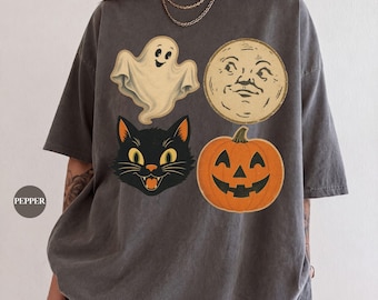 May include: A charcoal gray short-sleeve t-shirt featuring a graphic print of a cream-colored ghost, a beige moon with a face, a black cat head with yellow eyes, and an orange jack-o'-lantern. The word PEPPER is printed in a small circle.