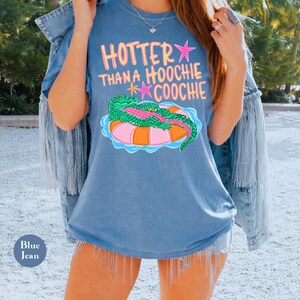 Puede incluir: Una camiseta azul con el texto "HOTTER THAN A HOOCHIE COOCHIE" en rosa y amarillo. La camiseta presenta un cocodrilo de dibujos animados en un flotador inflable rosa y naranja. Las palabras "Blue Jean" est&aacute;n impresas en un peque&ntilde;o c&iacute;rculo azul.