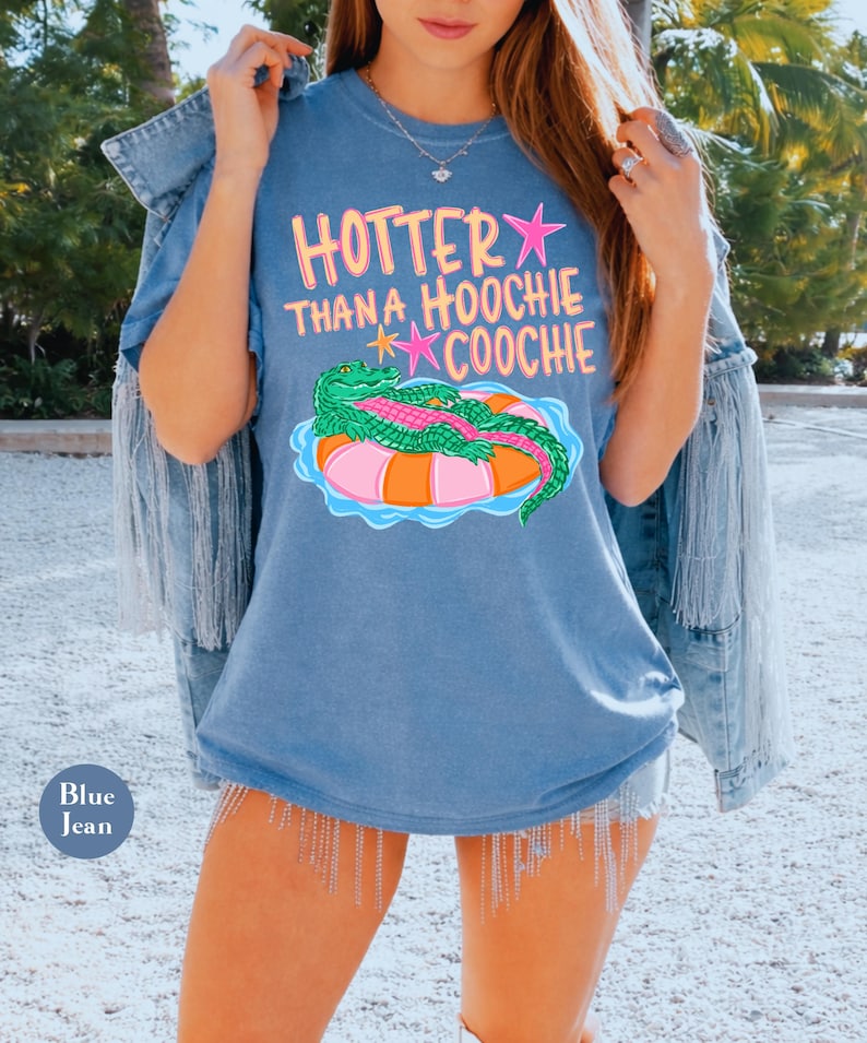 Puede incluir: Una camiseta azul con el texto "HOTTER THAN A HOOCHIE COOCHIE" en rosa y amarillo. La camiseta presenta un cocodrilo de dibujos animados en un flotador inflable rosa y naranja. Las palabras "Blue Jean" est&aacute;n impresas en un peque&ntilde;o c&iacute;rculo azul.