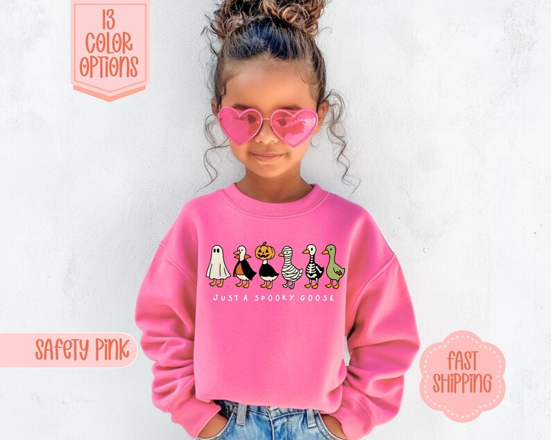 May include: A bright pink sweatshirt featuring the phrase "JUST A SPOOKY GOOSE" and a row of cartoon geese in Halloween costumes. The sweatshirt is worn by a child. The image also shows text that says "13 COLOR OPTIONS" and "SAFETY PINK".