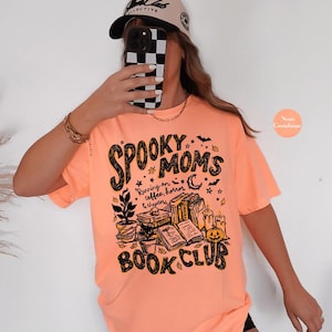 May include: A neon orange short-sleeve t-shirt featuring a black graphic print that reads Spooky Moms Book Club and Running on coffee, horror & chapters. The design includes illustrations of stacked books, a potted plant, candles, and a pumpkin.