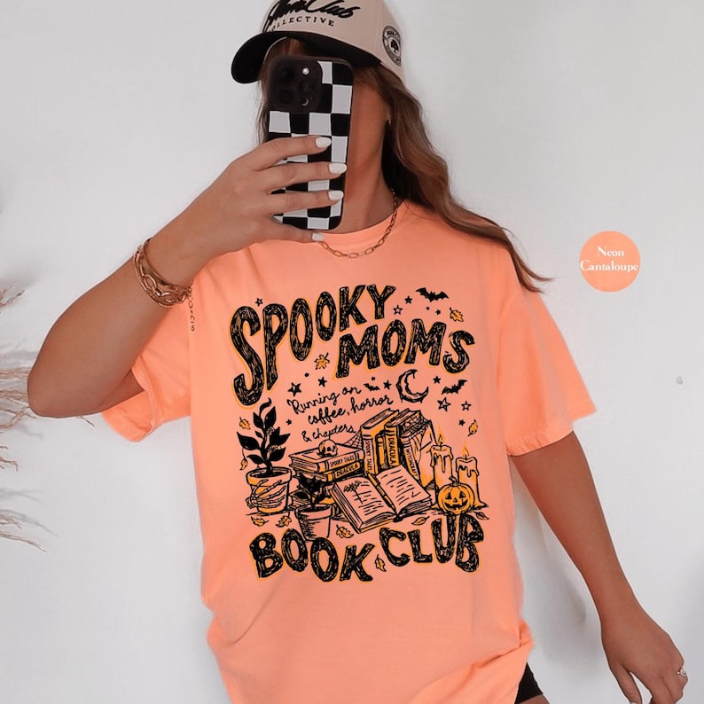 May include: A neon orange short-sleeve t-shirt featuring a black graphic print that reads Spooky Moms Book Club and Running on coffee, horror & chapters. The design includes illustrations of stacked books, a potted plant, candles, and a pumpkin.