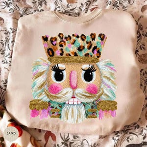 Puede incluir: Sudadera color arena con un diseño bordado de un cascanueces. El cascanueces tiene una corona con estampado de leopardo, ojos grandes, mejillas rosas y un bigote blanco. La palabra "SAND" está impresa en una etiqueta redonda en la esquina inferior izquierda.