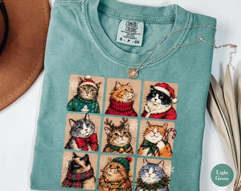 May include: A light green t-shirt featuring a graphic print of nine cats in a grid layout. Each cat is illustrated with festive holiday accessories including Santa hats, scarves, reindeer antlers, and Christmas lights. The shirt has a crew neckline.
