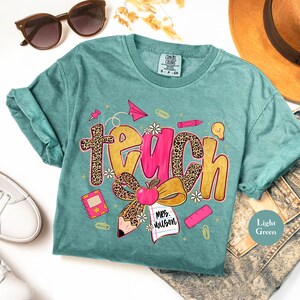May include: Light green t-shirt with the word "TEACH" in a playful design. The word "TEACH" has a leopard print pattern and pink accents. The shirt also features school-themed graphics like an apple, pencil, and paper airplane.