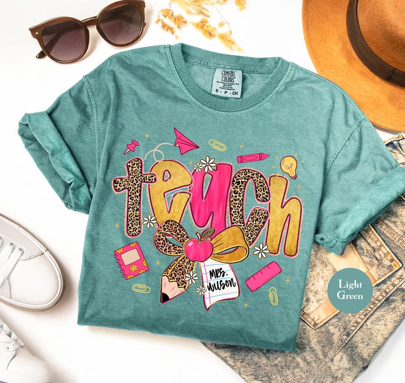 May include: Light green t-shirt with the word "TEACH" in a playful design. The word "TEACH" has a leopard print pattern and pink accents. The shirt also features school-themed graphics like an apple, pencil, and paper airplane.