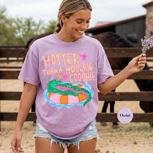 Puede incluir: Camiseta color orqu&iacute;dea con el texto "HOTTER THAN A HOOCHIE COOCHIE" en naranja y rosa. La camiseta presenta un gr&aacute;fico de un caim&aacute;n verde sobre un flotador inflable rosa y naranja. La modelo lleva pantalones cortos vaqueros.