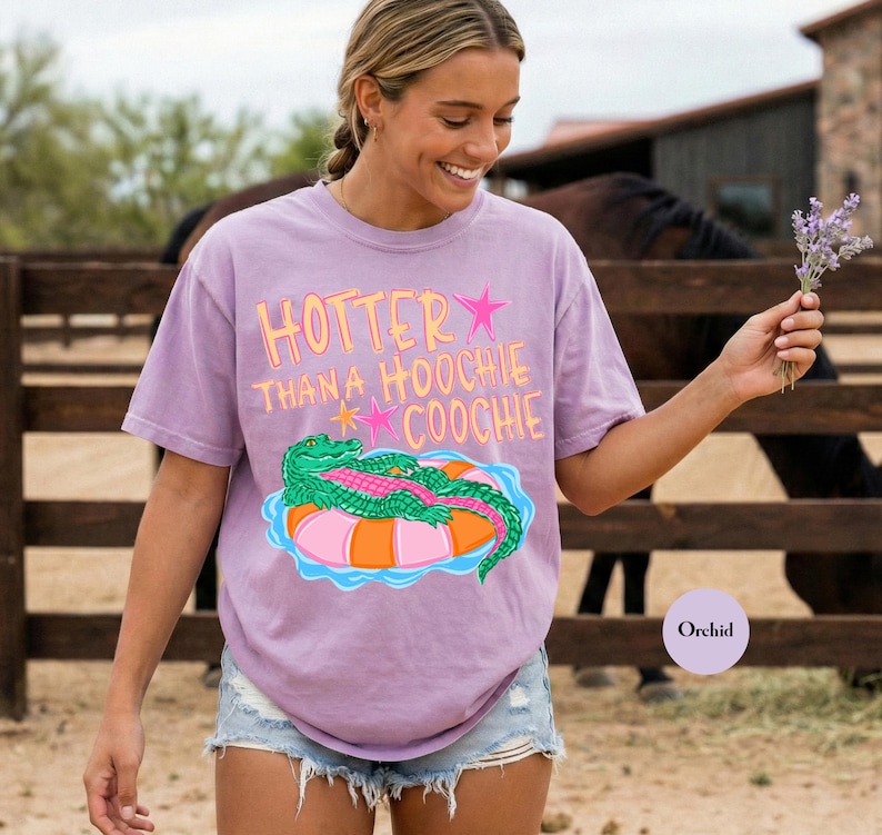 Puede incluir: Camiseta color orqu&iacute;dea con el texto "HOTTER THAN A HOOCHIE COOCHIE" en naranja y rosa. La camiseta presenta un gr&aacute;fico de un caim&aacute;n verde sobre un flotador inflable rosa y naranja. La modelo lleva pantalones cortos vaqueros.