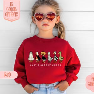 May include: A red sweatshirt with a Halloween-themed design. The design features a ghost, penguin, pumpkin, mummy, and goose. The text "JUST A SPOOKY GOOSE" is below the characters. The image also shows color options.