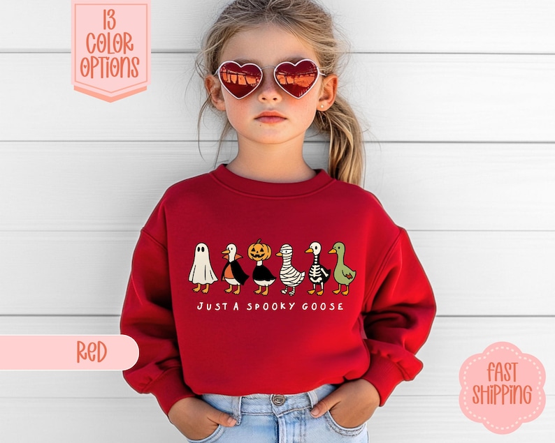 May include: A red sweatshirt with a Halloween-themed design. The design features a ghost, penguin, pumpkin, mummy, and goose. The text "JUST A SPOOKY GOOSE" is below the characters. The image also shows color options.