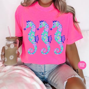 May include: Bright neon pink t-shirt featuring three seahorse designs. The seahorses are decorated with a floral pattern in shades of blue, green, and pink. The shirt has a crew neck and short sleeves. The words "Neon Pink" are in a pink circle.