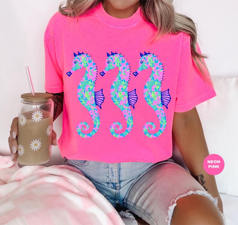 May include: Bright neon pink t-shirt featuring three seahorse designs. The seahorses are decorated with a floral pattern in shades of blue, green, and pink. The shirt has a crew neck and short sleeves. The words "Neon Pink" are in a pink circle.