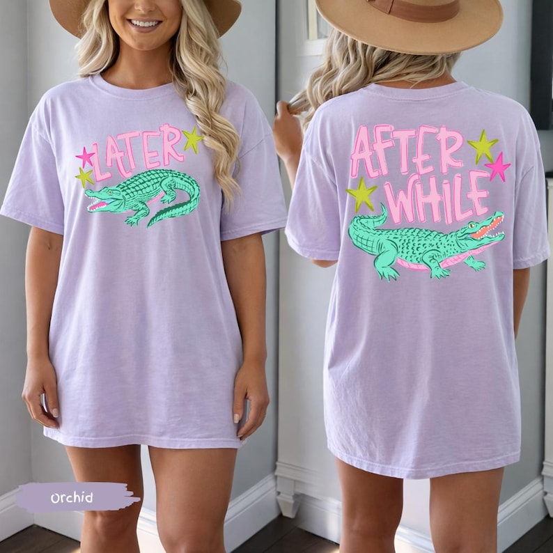 May include: Orchid-colored t-shirt with a front graphic of a green alligator and the word "LATER" in pink. The back of the shirt features the words "AFTER WHILE" and a matching alligator design. The shirt has short sleeves and a relaxed fit.