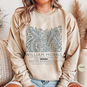Puede incluir: Sudadera color arena con un dise&ntilde;o floral en tonos azul-verdoso. El dise&ntilde;o incluye el texto "WILLIAM MORRIS COTTON PRINTS EXHIBITION" y detalles del evento. La sudadera es de cuello redondo.
