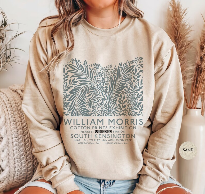 Puede incluir: Sudadera color arena con un dise&ntilde;o floral en tonos azul-verdoso. El dise&ntilde;o incluye el texto "WILLIAM MORRIS COTTON PRINTS EXHIBITION" y detalles del evento. La sudadera es de cuello redondo.
