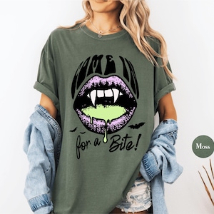 May include: A moss green short-sleeve t-shirt featuring a graphic of purple lips with white vampire fangs and dripping neon green liquid. The text "COME IN" appears above the lips and "for a Bite!" below, accompanied by two small black bats.