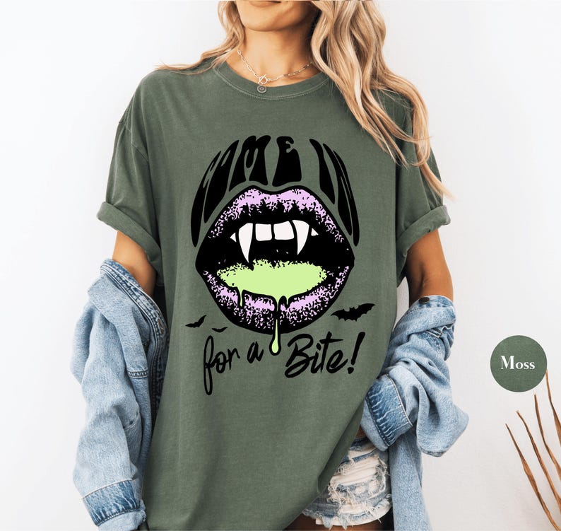 May include: A moss green short-sleeve t-shirt featuring a graphic of purple lips with white vampire fangs and dripping neon green liquid. The text "COME IN" appears above the lips and "for a Bite!" below, accompanied by two small black bats.