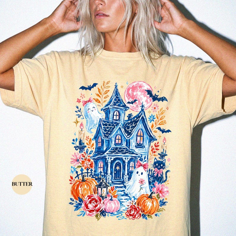 K&ouml;nnte beinhalten: Ein buttergelbes T-Shirt mit einem Grafikdruck eines blauen Spukhauses, zwei Geistern mit rosa Schleifen, orangefarbenen K&uuml;rbissen, roten Blumen, einer schwarzen Katze, Flederm&auml;usen und einem rosa Mond sowie Kerzen und floralen Akzenten.