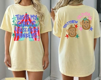 May include: A pale yellow t-shirt featuring a graphic of a circus tent with the text "THIS IS MY CIRCUS & THESE ARE MY MONKEYS" on the front, and two monkey faces with the names "MATTHEW" and "OLIVIA" surrounded by colorful stars on the back.