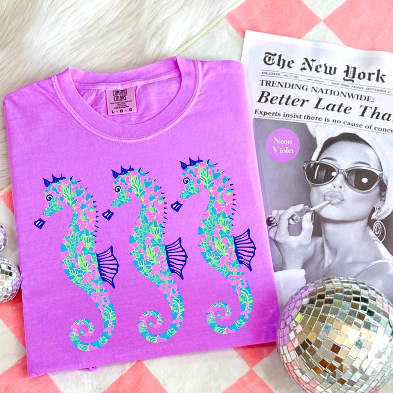 May include: A lilac Comfort Colors t-shirt featuring three seahorse designs. The seahorses are decorated with a floral pattern in shades of pink, green, and blue. A newspaper with the headline "Better Late Than" and a disco ball are also in the image.
