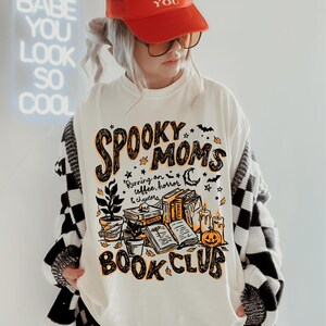 May include: An off-white t-shirt featuring a graphic print with the text "SPOOKY MOMS BOOK CLUB" and "Running on coffee, horror & chapters". The design includes illustrations of stacked books, a potted plant, candles, and a jack-o'-lantern.