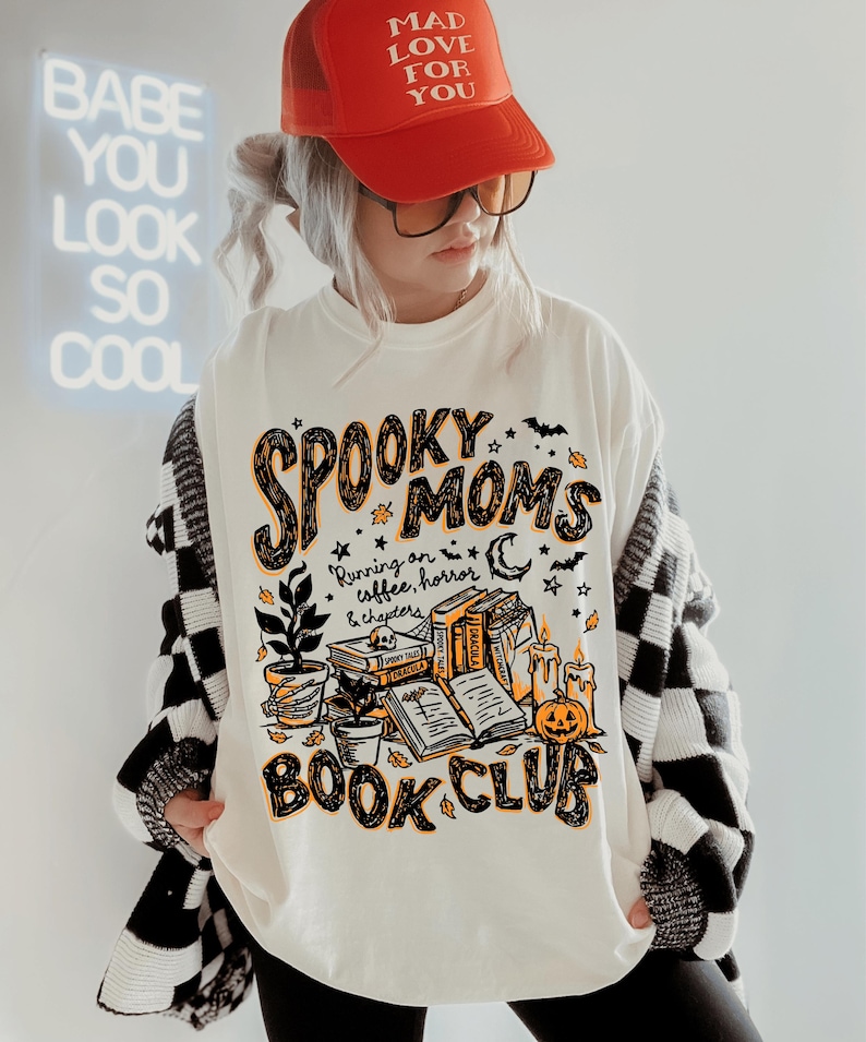 May include: An off-white t-shirt featuring a graphic print with the text "SPOOKY MOMS BOOK CLUB" and "Running on coffee, horror & chapters". The design includes illustrations of stacked books, a potted plant, candles, and a jack-o'-lantern.