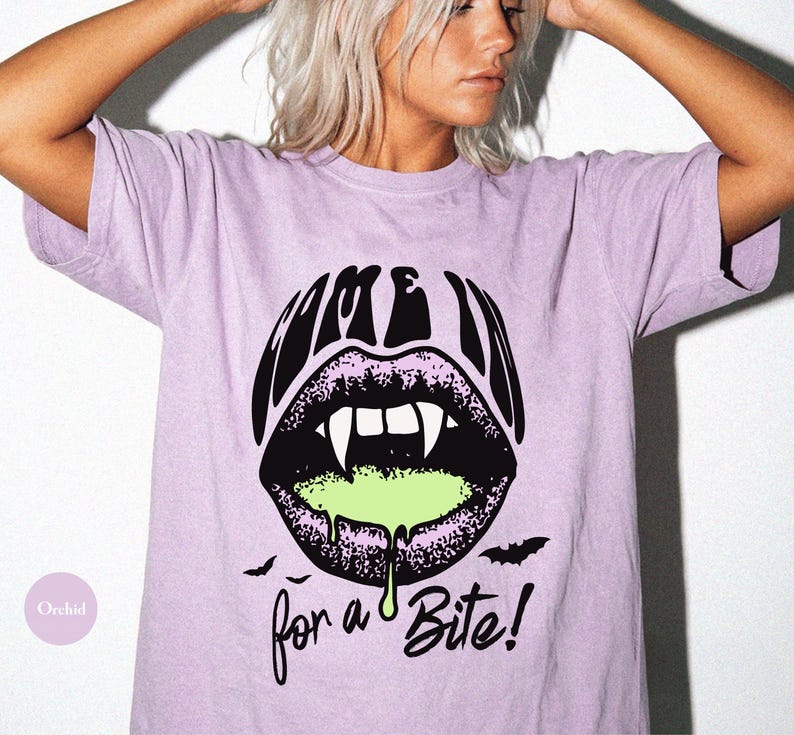 May include: A light purple t-shirt featuring a graphic of black lips with white vampire fangs and dripping neon green liquid. The text above the lips reads "COME IN" and the text below reads "for a Bite!" with two small black bats on the sides.
