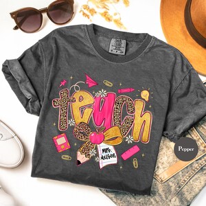 May include: Dark gray t-shirt with the word "Teach" in gold and pink, featuring a leopard print design. The shirt includes school-themed graphics like an apple, pencil, and paper. The shirt is a Comfort Colors brand.