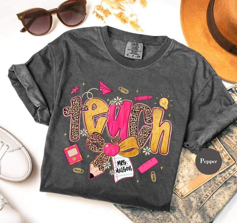 May include: Dark gray t-shirt with the word "Teach" in gold and pink, featuring a leopard print design. The shirt includes school-themed graphics like an apple, pencil, and paper. The shirt is a Comfort Colors brand.
