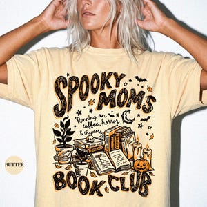 May include: A butter-colored t-shirt featuring a graphic print with the text SPOOKY MOMS BOOK CLUB and Running on coffee, horror & chapters. The design includes illustrations of stacked books, a skull, a potted plant, candles, and a pumpkin.
