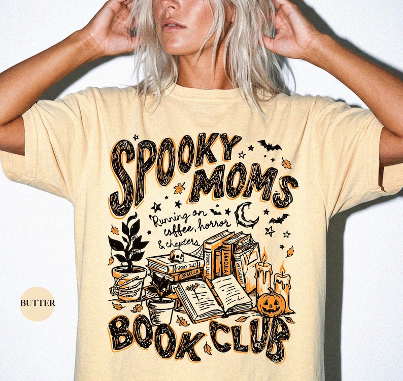 May include: A butter-colored t-shirt featuring a graphic print with the text SPOOKY MOMS BOOK CLUB and Running on coffee, horror & chapters. The design includes illustrations of stacked books, a skull, a potted plant, candles, and a pumpkin.