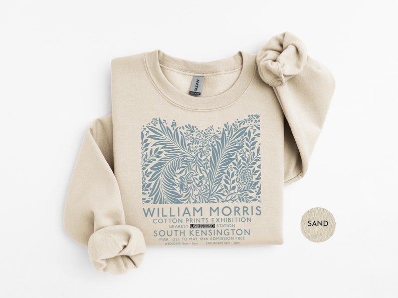 Puede incluir: Una sudadera de color arena con un dise&ntilde;o floral azul y el texto "WILLIAM MORRIS COTTON PRINTS EXHIBITION" y detalles de ubicaci&oacute;n en South Kensington. La sudadera tiene cuello redondo y mangas largas.