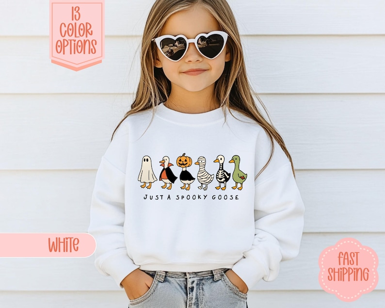 May include: White sweatshirt with a Halloween-themed design. It features cartoon geese in costumes and the phrase "JUST A SPOOKY GOOSE". The image also includes text that says "13 COLOR OPTIONS" and "WHITE".