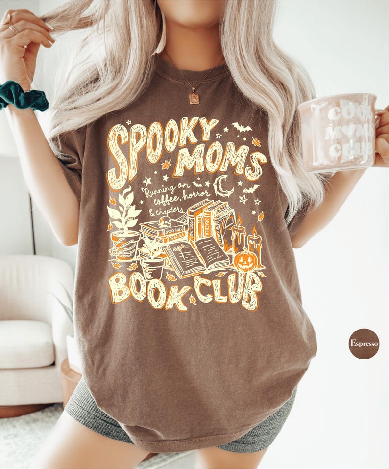 May include: A brown short-sleeve t-shirt featuring a graphic print with the text "SPOOKY MOMS BOOK CLUB" and "Running on coffee, horror & chapters". The design includes illustrations of stacked books, a pumpkin, candles, and stars.