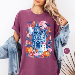 K&ouml;nnte beinhalten: Ein beerenfarbenes Kurzarm-T-Shirt mit einem Grafikdruck eines blauen Spukhauses, Geistern mit rosa Schleifen, orangefarbenen K&uuml;rbissen, roten Rosen, einer schwarzen Katze, Flederm&auml;usen und einem Vollmond. Dazu eine blaue Jeansjacke &uuml;ber den Schultern.