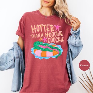 Puede incluir: Camiseta color carmes&iacute; con la frase "Hotter Than A Hoochie Coochie" en rosa y amarillo. El dise&ntilde;o presenta un caim&aacute;n verde sobre un flotador inflable rosa y naranja. La camiseta tiene mangas cortas.