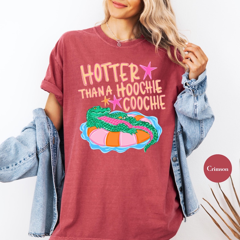 Puede incluir: Camiseta color carmes&iacute; con la frase "Hotter Than A Hoochie Coochie" en rosa y amarillo. El dise&ntilde;o presenta un caim&aacute;n verde sobre un flotador inflable rosa y naranja. La camiseta tiene mangas cortas.