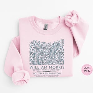 Puede incluir: Sudadera rosa claro con un dise&ntilde;o floral gris y el texto "WILLIAM MORRIS COTTON PRINTS EXHIBITION SOUTH KENSINGTON". El dise&ntilde;o incluye elementos bot&aacute;nicos y texto. La sudadera es de color rosa claro.