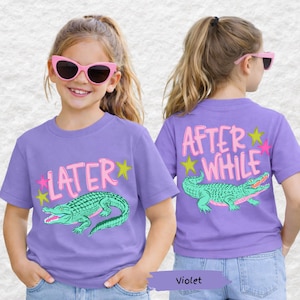 May include: Two children wearing violet t-shirts with a green alligator graphic. The front of the shirt says "LATER" in pink letters with yellow and pink stars. The back of the shirt says "AFTER WHILE" with the same alligator graphic.