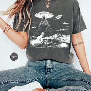 Może przedstawiać: Szary t-shirt z białym nadrukiem przedstawiającym kota patrzącego w g&oacute;rę na UFO. UFO emituje wiązki światła, a mały statek kosmiczny leci w pobliżu. Nadruk zawiera r&oacute;wnież pasmo g&oacute;rskie i chmury.