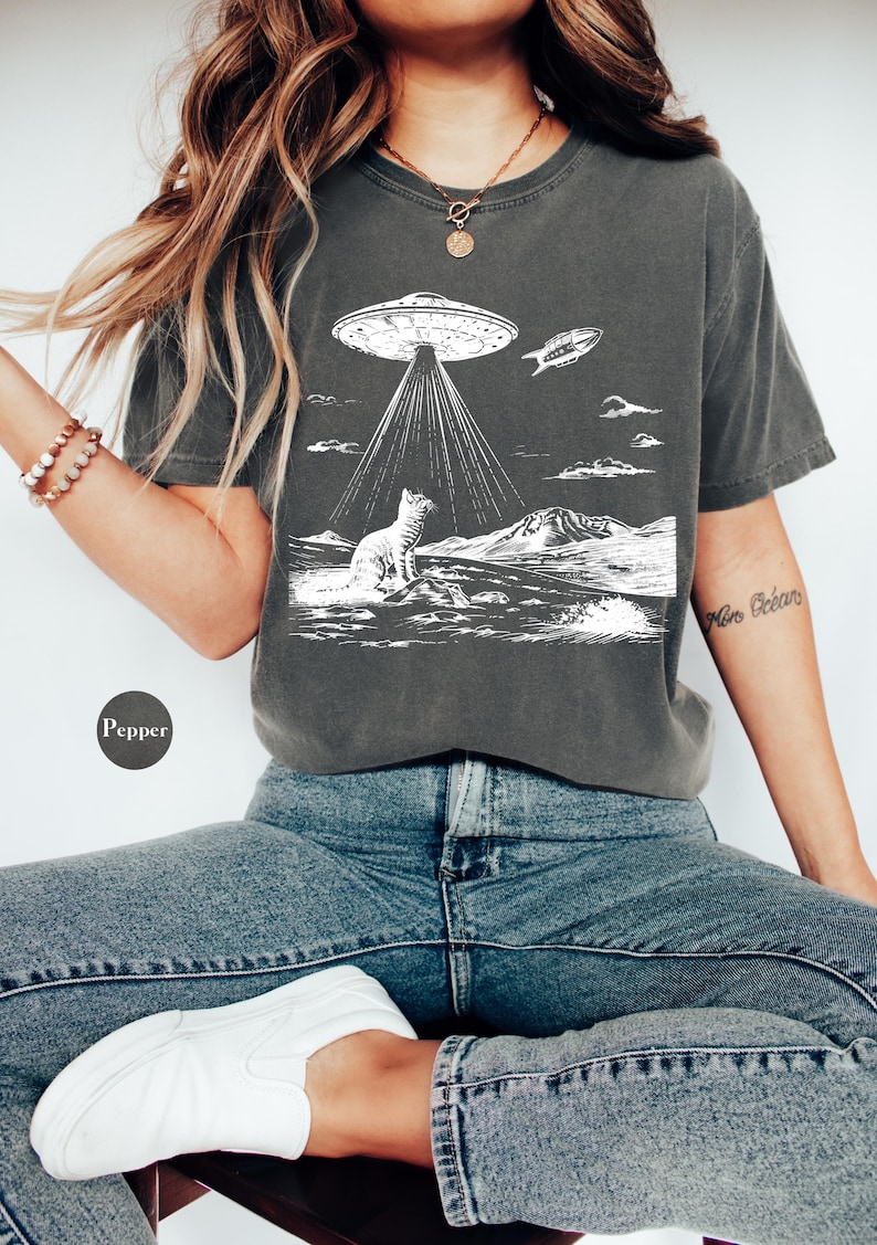 Może przedstawiać: Szary t-shirt z białym nadrukiem przedstawiającym kota patrzącego w g&oacute;rę na UFO. UFO emituje wiązki światła, a mały statek kosmiczny leci w pobliżu. Nadruk zawiera r&oacute;wnież pasmo g&oacute;rskie i chmury.