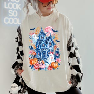 K&ouml;nnte beinhalten: Ein elfenbeinfarbenes T-Shirt mit einem farbenfrohen Grafikdruck eines blauen Spukhauses, zwei wei&szlig;en Geistern, orangefarbenen K&uuml;rbissen, rosa Blumen, einer schwarzen Katze und Flederm&auml;usen unter einem rosa Mond mit floralen Akzenten.