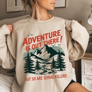 K&ouml;nnte beinhalten: Beiges Sweatshirt mit dem roten Text "ADVENTURE IS OUT THERE!" &uuml;ber einer Berg- und Waldgrafik. Darunter steht "BUT SO ARE SERIAL KILLERS". Das Sweatshirt hat lange &Auml;rmel.