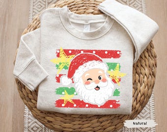 May include: A natural-colored crewneck sweatshirt featuring a graphic print of a winking Santa Claus face. The design includes horizontal red and green distressed stripes with white polka dots and three yellow hand-drawn style stars.
