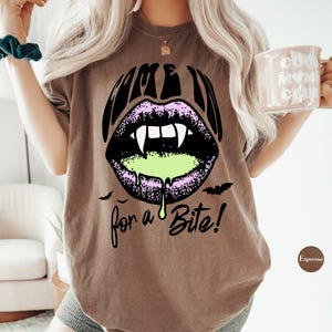 May include: A brown short-sleeve t-shirt featuring a graphic of purple lips with white fangs and green slime dripping from the mouth. The text "COME IN" is printed above the lips and "for a Bite!" is printed below, accompanied by two small black bats.